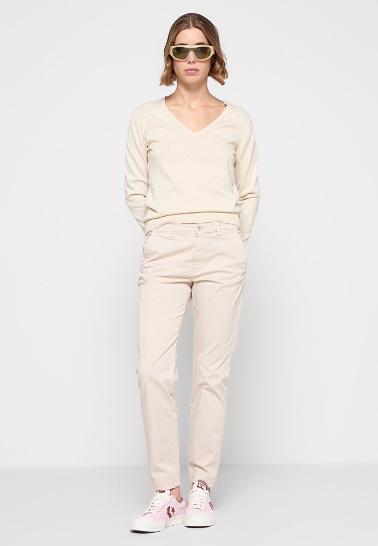 Beige v-hals trui gecombineerd met lichtbeige op maat gemaakte broek en witte sneakers met roze accenten. Model draagt getinte zonnebril.