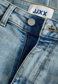 Jasnoniebieskie jeansy z denimu z zamkiem błyskawicznym, metalowym guzikiem i metką marki "JJXX" w pogrubionym niebieskim napisie na pasie.