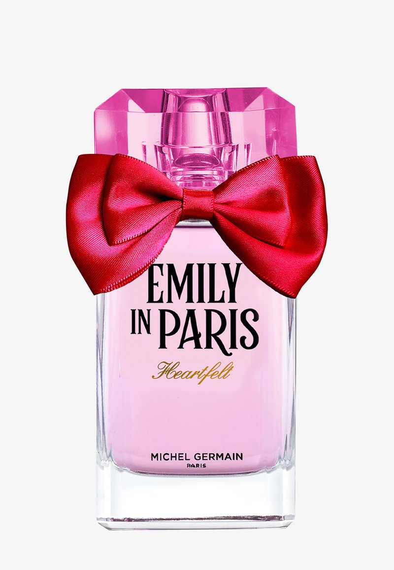 Flacon de parfum rose clair étiqueté Emily in Paris Heartfelt par Michel Germain avec un grand nœud en satin rouge attaché autour du col.