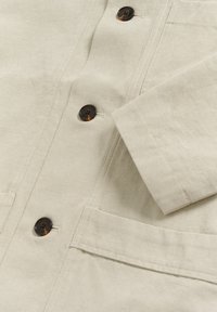 Beżowa tkanina kurtki z guzikiem o wzorze muszli żółwia, widocznymi dziurkami na guziki, złożonym rękawem i przeszyciami przy przedniej kieszeni.