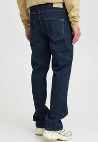 Tamsiai mėlyni džinsai tiesiu kirpimu, sukurtos su dviem užpakalinėmis kišenėmis ir smėlio spalvos juosmeniu. Dėvimi su šviesiomis sportinėmis batelėmis.