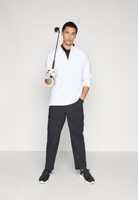Λευκό φούτερ με φερμουάρ, μαύρο πουκάμισο με γιακά, σκούρο παντελόνι cargo και μαύρα αθλητικά παπούτσια. Κρατάει μπαστούνι γκολφ με λευκό γάντι. Απλό φόντο.