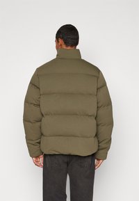 Giacca puffer verde oliva con colletto alto, design trapuntato e imbottitura spessa. Abbinata a jeans scuri, vista da dietro.