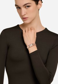Orologio da polso in oro rosa con quadrante rettangolare, dial argento e bracciale metallico a maglie. Indossato su una mano con pelle chiara, su uno sfondo neutro.