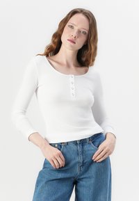 Hvit ribbet langermet topp med firkantet utringning, med fem metalliske knapper foran, kombinert med blå jeans.
