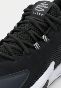 Black athletic shoe featuring a mesh and synthetic upper, textured finish, contrasting white accents, and a logo tag labeled "Curry."