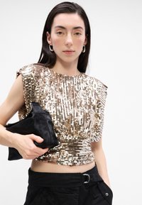 Top strălucitor din paiete aurii și argintii, cu un design ajustat. Modelul ține în mână o geantă clutch din blană sintetică neagră. Este asortat cu pantaloni cargo negri.