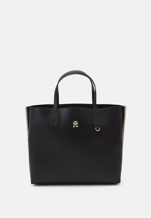 Sac cabas en cuir noir avec deux poignées, petit logo doré et détail anneau à l'avant, fond uni clair.