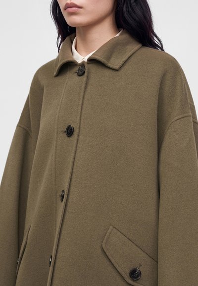 Mujer con un abrigo verde oliva con botones, cuello ancho y bolsillos con solapa sobre una camisa blanca con fondo liso.
