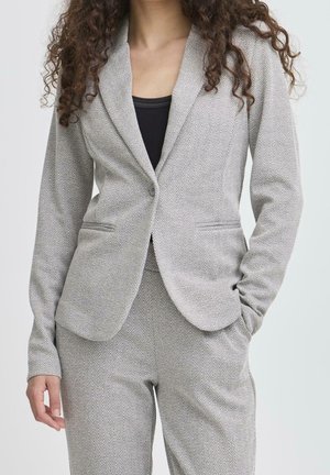 Femme portant un blazer à chevrons gris clair et un pantalon assorti avec un haut noir en dessous, debout avec une main dans la poche.