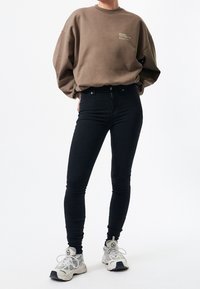 Hnědá oversized mikina s grafickým potiskem, spárovaná s černými skinny džíny a bílými teniskami se šedými detaily a tkaničkami.