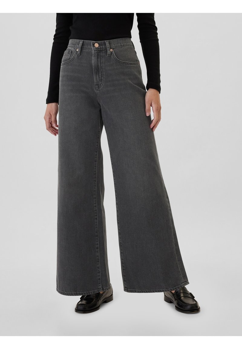 Jean en denim gris à jambes larges avec une taille haute, poches avant et fermeture à bouton. Texture douce et coupe ajustée. Des chaussures noires complètent le look.