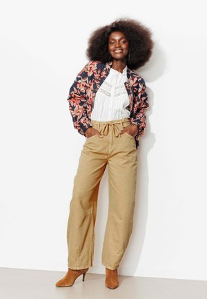 Femme souriante avec une coiffure afro naturelle porte une veste bomber fleurie, un chemisier blanc, un pantalon beige taille haute et des bottines à talons marron, debout en position détendue.