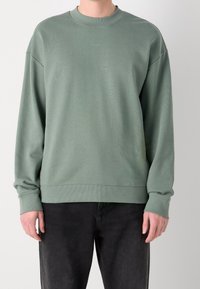 Uomo che indossa una felpa HUGO verde tenue con polsini e fondo a costine, abbinata a jeans neri, su uno sfondo chiaro e uniforme.