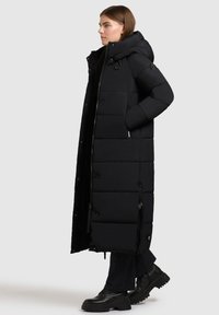 Cappotto lungo imbottito nero con cappuccio, caratterizzato da un design trapuntato, bottoni a pressione, dettagli con zip e tasche laterali. Tessuto liscio e isolato.