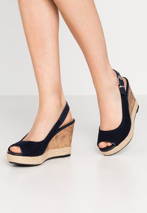 Piernas femeninas con sandalias negras de cuña con puntera abierta, tacones de corcho y tiras en el tobillo, de pie sobre un suelo claro.