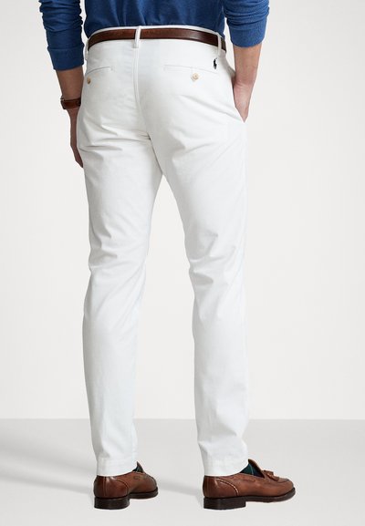 Valkoiset puuvillahousut suoralla leikkauksella, joissa on kaksi takataskua nappikiinnityksellä ja vyötärö, jota korostaa ruskea nahkavyö.