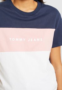 Puuvillane T-särk sinise, roosa ja valge värviliste plokkidega. Sellel on roosa osas valgete tähtedega tekst "TOMMY JEANS". Lühikesed varrukad.