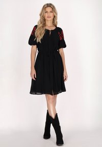 Schwarzes Kleid mit Puffärmeln, das über rote Blumenstickereien an den Schultern, einer taillierten Taille und einer leichten, fließenden Textur verfügt. Kombiniert mit schwarzen Stiefeln.
