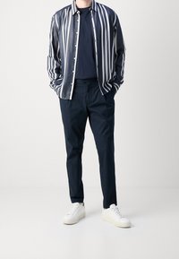 Skjorta med knappar i marinblå och vita vertikala ränder, kombinerad med mörka byxor och vita sneakers, vilket visar en avslappnad look och en ledig passform.