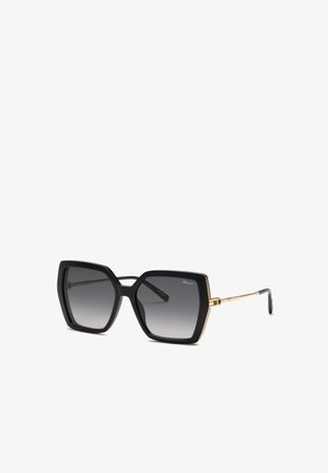 Lunettes de soleil carrées noires surdimensionnées avec des verres gris dégradés et des fines branches dorées inclinées sur un fond blanc.