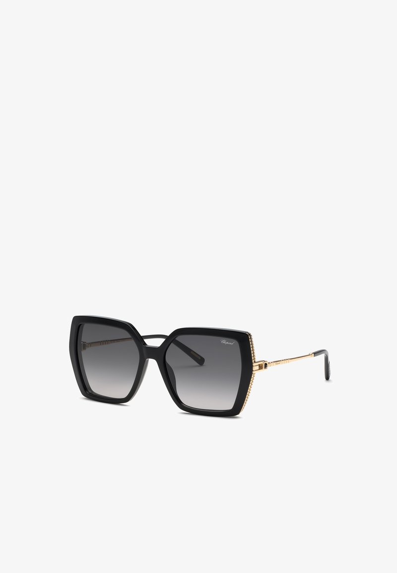 Lunettes de soleil carrées noires surdimensionnées avec des verres gris dégradés et des fines branches dorées inclinées sur un fond blanc.