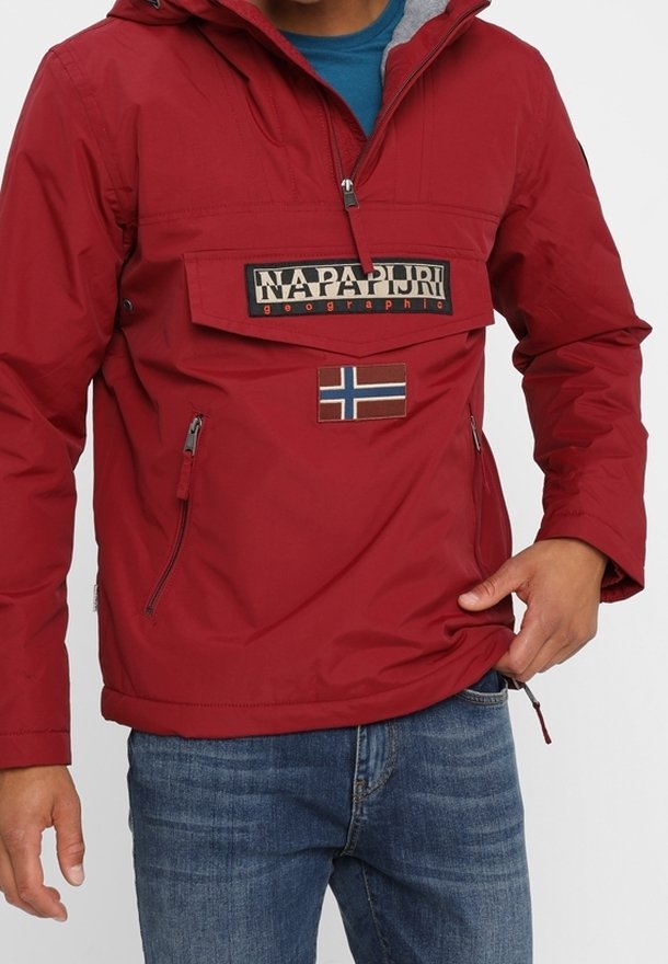 Veste rouge Napapijri avec poche avant, écusson du drapeau norvégien et poches latérales zippées portée avec un jean bleu.