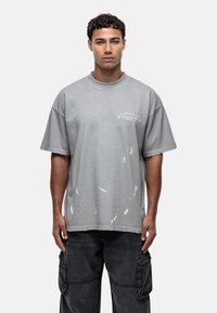 T-shirt oversize gris en coton avec un col rond, présentant des éclaboussures de peinture blanche et un petit graphique sur la poitrine.