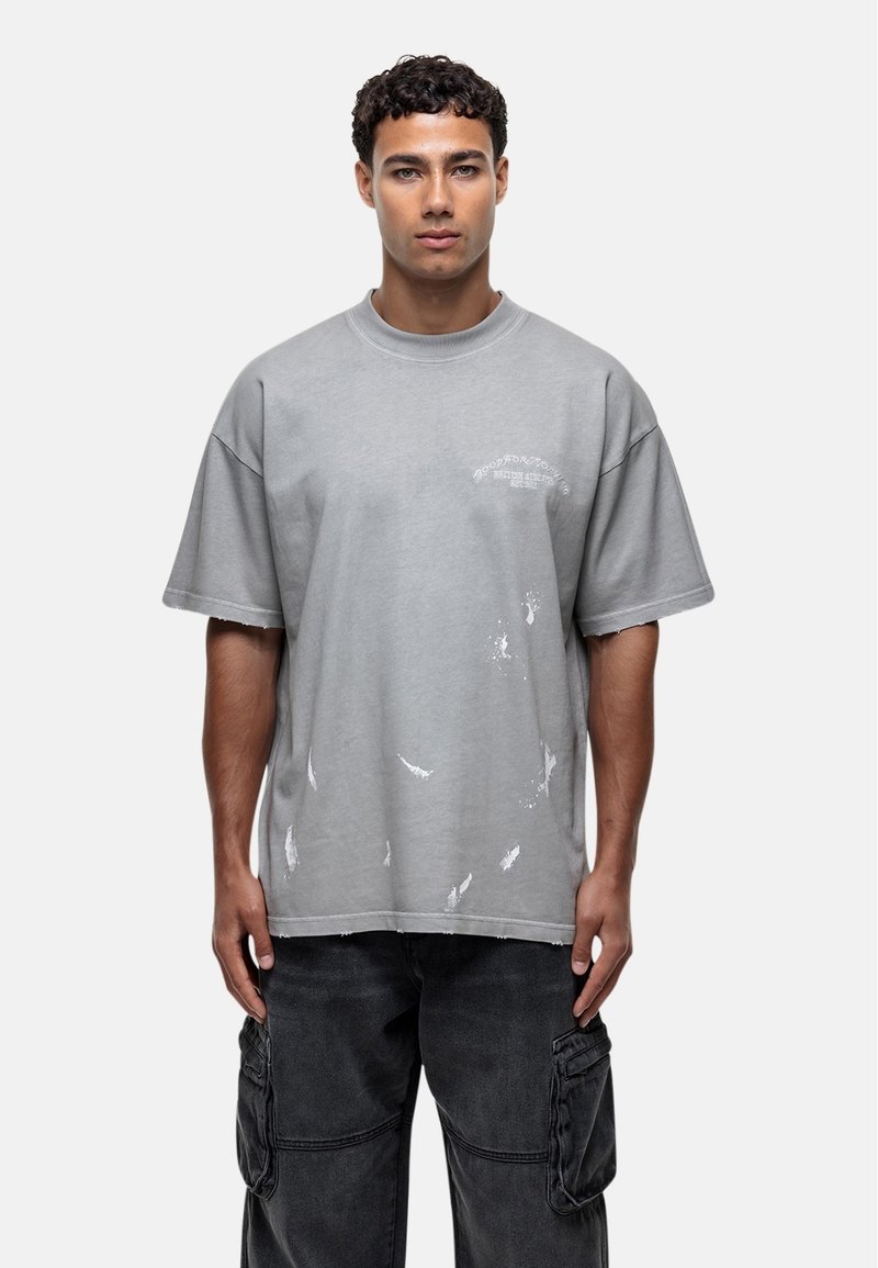 T-shirt oversize gris en coton avec un col rond, présentant des éclaboussures de peinture blanche et un petit graphique sur la poitrine.