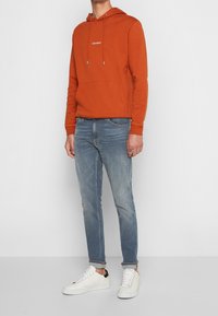 Orange bomullshuvtröja med en framficka och snörning, tillsammans med ljusblå denimjeans och vita sneakers. Enkel design, avslappnad passform.