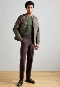 Casaco bomber acolchoado caqui sobre um suéter verde escuro, combinado com calças bordô escuras e mocassins castanhos, sobre um chão de madeira.