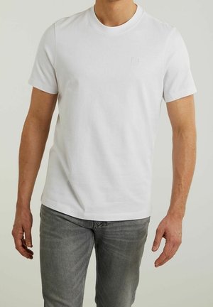 Homme portant un T-shirt blanc uni à manches courtes et un jean gris, debout devant un fond clair uni.