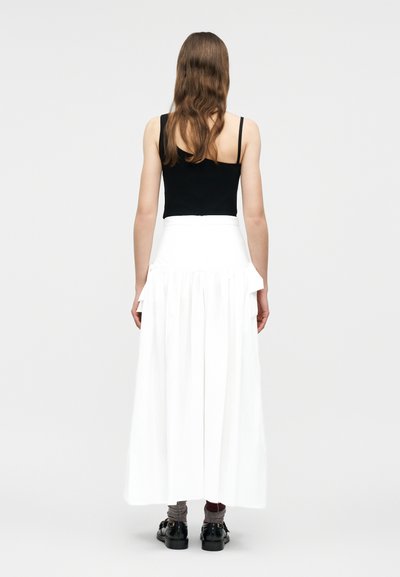 Γυναίκα με μακριά καστανά μαλλιά, φορούσε μαύρο αμάνικο τοπ, λευκή φούστα μέχρι τον αστράγαλο με πιέτες και ψηλή μέση, γκρι κάλτσες και μαύρα παπούτσια, στέκεται με την πλάτη στραμμένη.