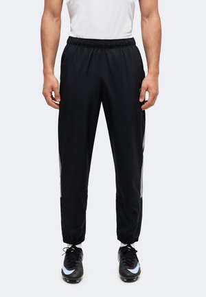 Homme portant un pantalon de sport noir avec des poignets élastiques, des bandes blanches sur les côtés et des chaussures de football noires avec des accents bleu clair.