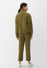 Frau steht mit dem Rücken zur Kamera, trägt eine olivgrüne Jacke und Hose sowie weiße Sneakers, vor einem schlichten weißen Hintergrund.