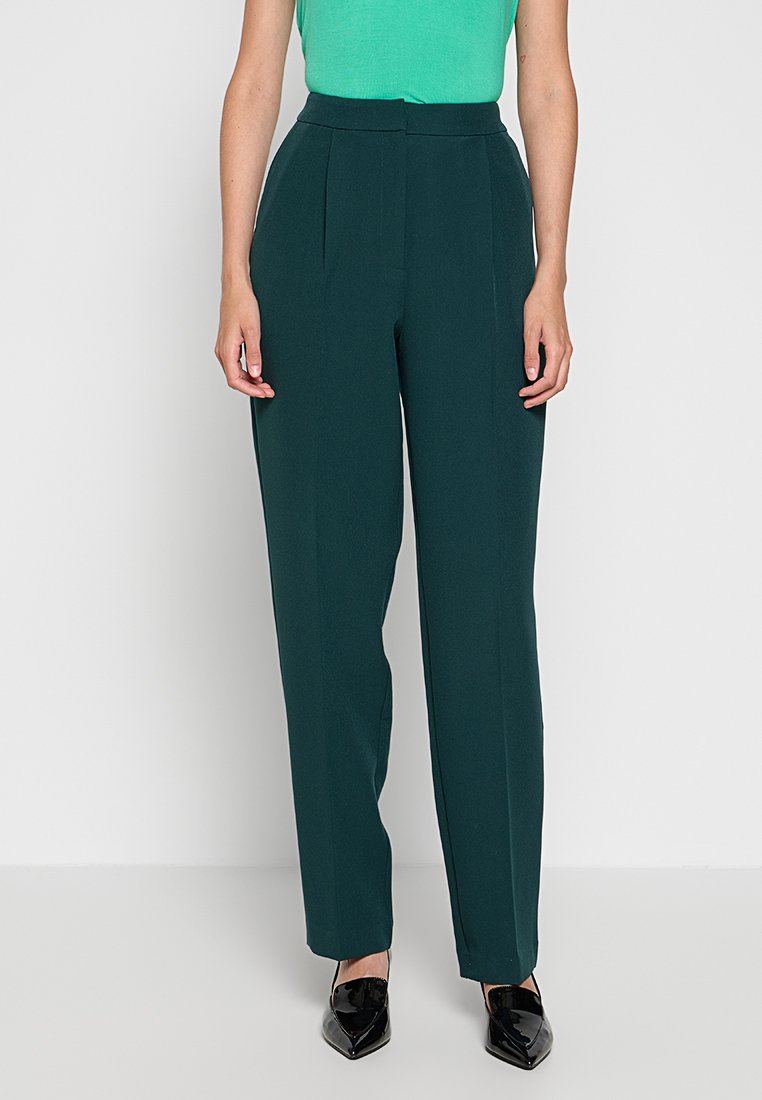 Vrouw in donkergroene, op maat gemaakte broek met hoge taille, zwarte puntige schoenen en een felgroene mouwloze top die is ingestopt.