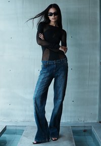 Mujer con cabello largo y oscuro, que lleva gafas de sol, top negro transparente, vaqueros anchos azules y zapatos abiertos, se sostiene con confianza junto a una pared de hormigón.
