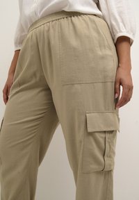 Pantaloni cargo color khaki realizzati in tessuto leggero. Vita elastica, tasche laterali e tasche in stile cargo su ogni gamba. Dettagli di cucitura minimi.