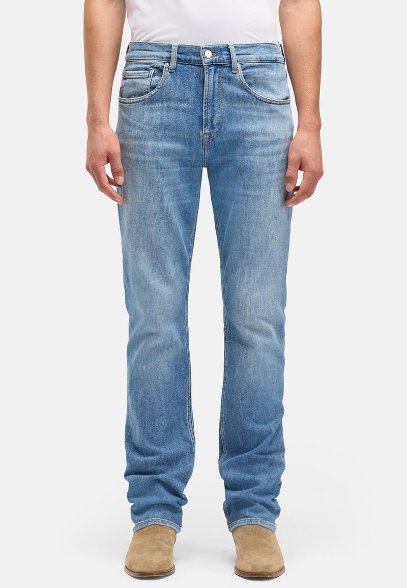 Homme portant un jean bleu clair à jambes droites et des bottes en daim marron clair, debout avec les bras détendus le long du corps devant un fond blanc.