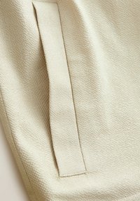 Tejido beige texturizado con una costura vertical prominente y un detalle de correa plana tejida. La superficie tiene un sutil patrón acanalado.