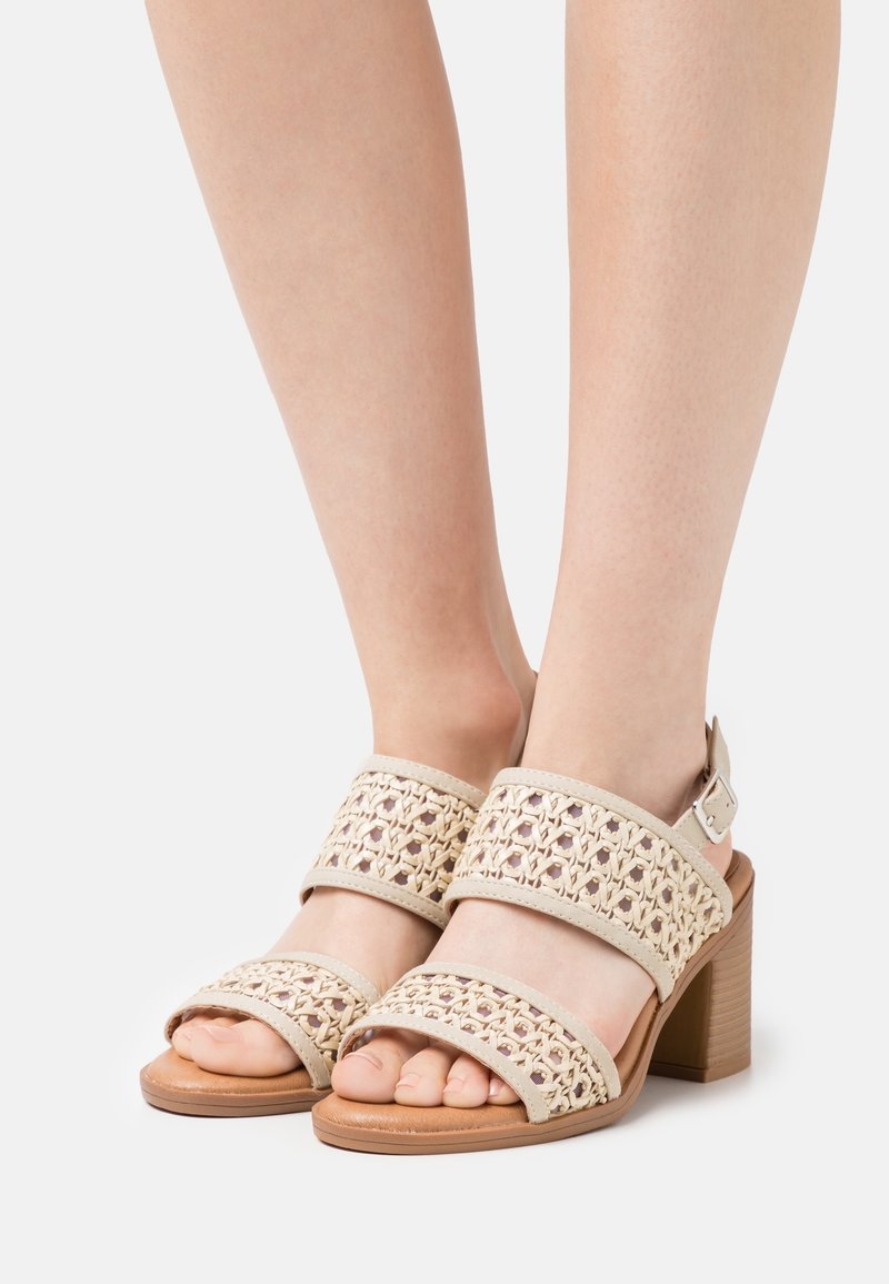 Pieds portant des sandales beiges tissées à bouts ouverts avec talons blocs et brides de cheville sur fond blanc.