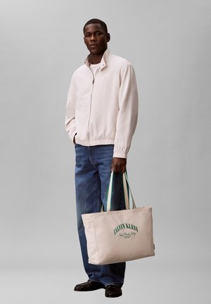 Bărbat tânăr purtând o jachetă deschisă pală și blugi, ținând o geantă tote Calvin Klein bej cu mânere cu dungi verzi.