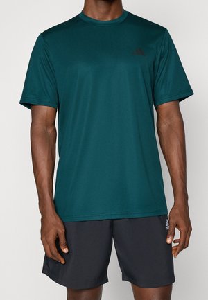 Man wearing a dark green short-sleeve athletic shirt and black sports shorts, standing against a plain background.