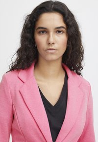 Blazer rosa con rever a contrasto, tessuto testurizzato e vestibilità sartoriale, indossato sopra una maglietta nera scollo a V. Sullo sfondo c'è un bianco uniforme.