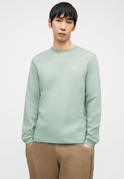 Άνδρας με μαύρα μαλλιά, που φοράει ανοιχτοπράσινο μακρυμάνικο πουκάμισο και καφέ παντελόνι, στέκεται με τα χέρια στις τσέπες μπροστά σε απλό φόντο.