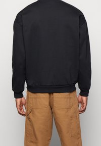 Sudadera negra con puños y dobladillo de canalé, hecha de una tela suave, usada sobre pantalones cargo color beige. Presenta un diseño clásico de cuello redondo.