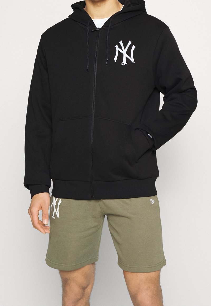 Felpa nera con zip e logo bianco NY sul lato sinistro del petto, abbinata a pantaloni corti verde oliva. Il materiale sembra morbido, con tasche anteriori.