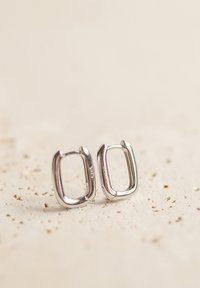 Srebrne kwadratowe kolczyki o zaokrąglonych krawędziach; gładka tekstura; minimalistyczny design; widoczne grawerowanie marki od wewnętrznej strony.