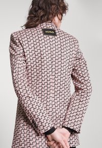 Blazer de la marca Hugo en un patrón geométrico rosa, con una etiqueta negra, corte estructurado y tejido texturizado. Se muestra la vista trasera.