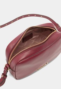 Burgundská kožená crossbody taška se zlatými detaily, zipovým uzavřením a texturovaným povrchem. Podšité interiér obsahuje vzor s logem.